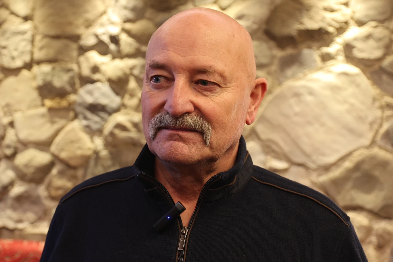 Starszy mężczyzna z łysą głową i siwym wąsem stoi na tle kamiennej ściany, ubrany w ciemny sweter z zamkiem, z mikrofonem przypiętym do kołnierza.