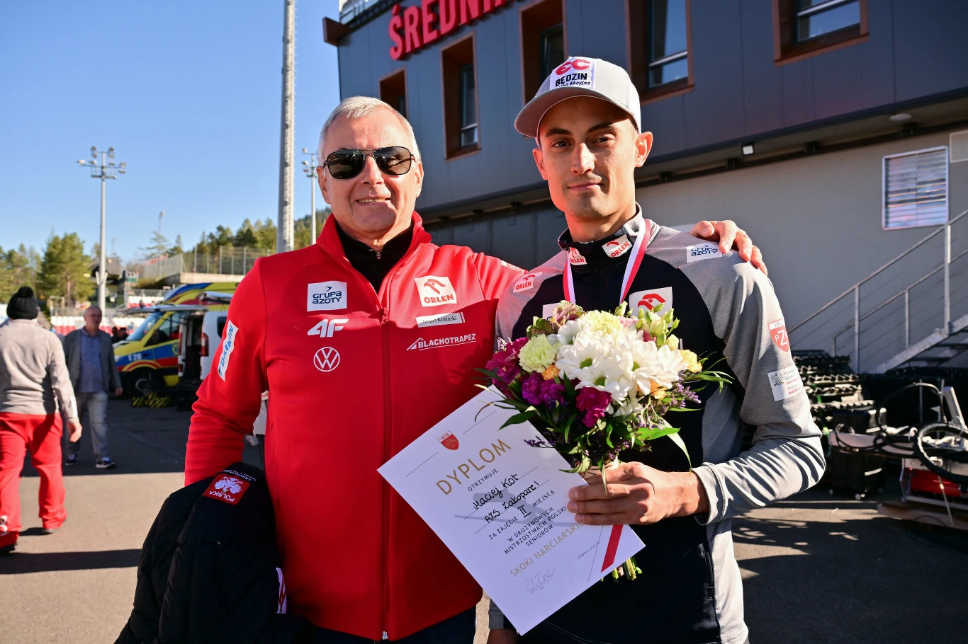 Dwóch mężczyzn, jeden trzyma bukiet kwiatów i dyplom, obaj uśmiechnięci, ubrani w sportowe stroje z logo, stoją na tle budynku z napisem 'ŚREDNIA'. Słoneczna pogoda, w tle inni ludzie i zaparkowane pojazdy.