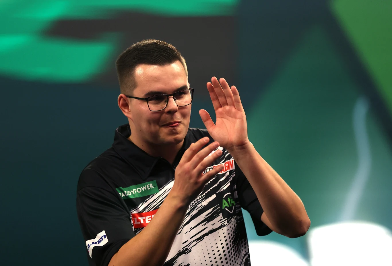 Gian van Veen Młody mężczyzna w okularach w sportowej koszulce z nadrukami sponsorów, stoi na tle zielonej scenografii i wykonuje gest dłońmi, jakby oklaskiwał lub dziękował publiczności. Wygląda na skupionego i zadowolonego.