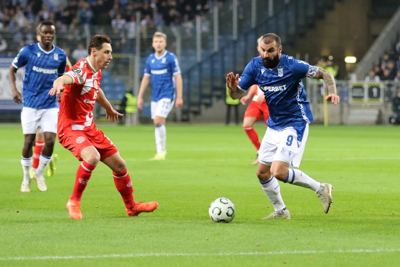 Lech Poznań zremisował z Mainz 1:1 Dwaj piłkarze konkurują o piłkę na tle stadionu pełnego widzów, zawodnik w niebieskiej koszulce prowadzi piłkę, a gracz w czerwonym stroju próbuje ją przejąć, pozostali zawodnicy obserwują sytuację.