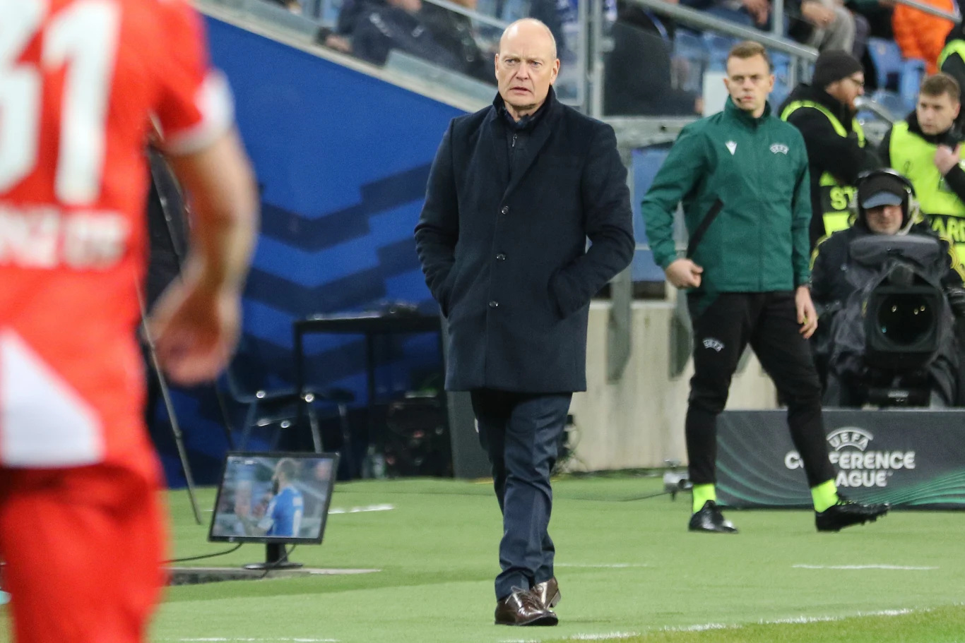 Lech Poznań zremisował z Mainz 1:1 Trener w eleganckim płaszczu stoi przy linii bocznej boiska podczas meczu piłki nożnej. W tle widoczni są zawodnicy, sędziowie techniczni i fragment trybun stadionu. Po lewej stronie widoczny piłkarz w czerwonym stroju z numerem 31.