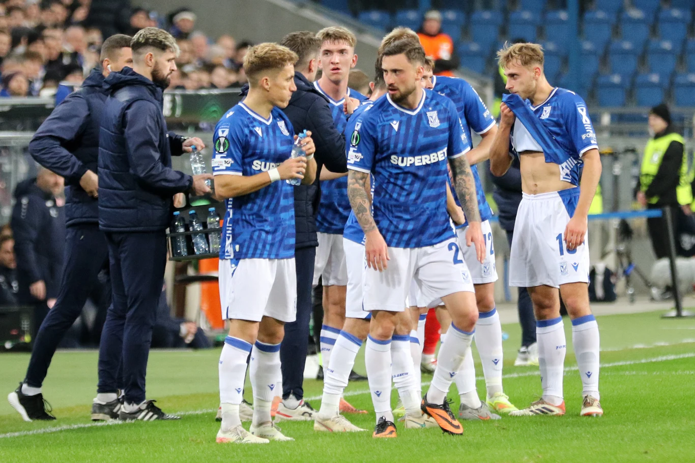 Grupa piłkarzy w niebieskich koszulkach konsultuje się na boisku podczas przerwy w meczu, w tle widoczne są trybuny z kibicami oraz sztab szkoleniowy.