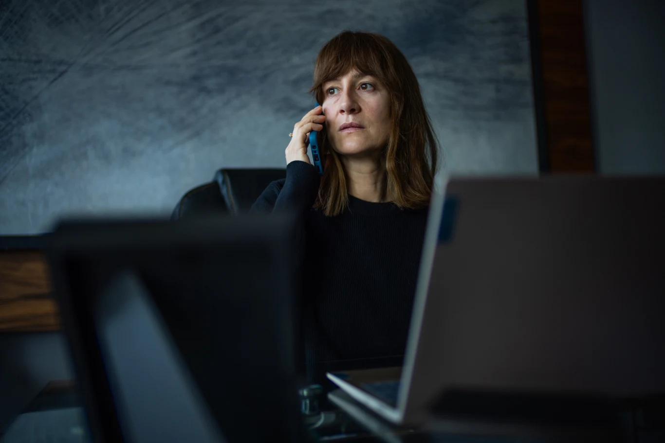 Kobieta siedząca przy biurku rozmawia przez telefon, na pierwszym planie widoczny ekran laptopa, wnętrze biura w tle.