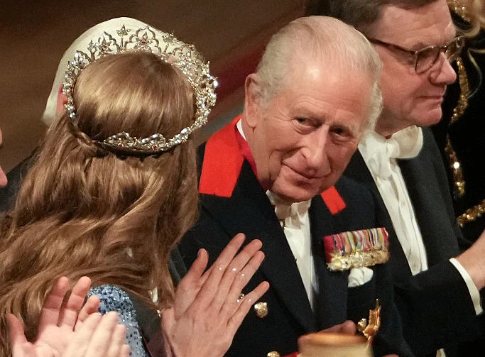 Starszy mężczyzna w mundurze wojskowym z odznaczeniami siedzi w sali podczas oficjalnej uroczystości, obok kobieta z diademem na głowie oraz mężczyzna w okularach i smokingu. Postacie ubrane są bardzo elegancko i biorą udział w ważnym wydarzeniu.