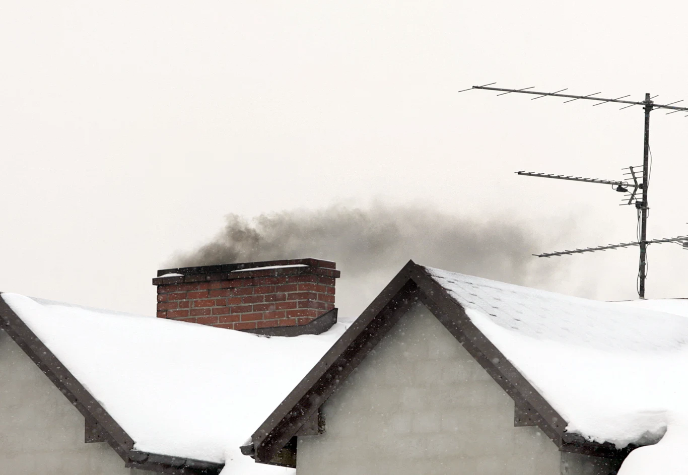 W czasie zimy coraz więcej kominów intensywnie dymi Dach domu pokryty śniegiem, z komina unosi się ciemny dym, obok zamontowane są anteny telewizyjne.