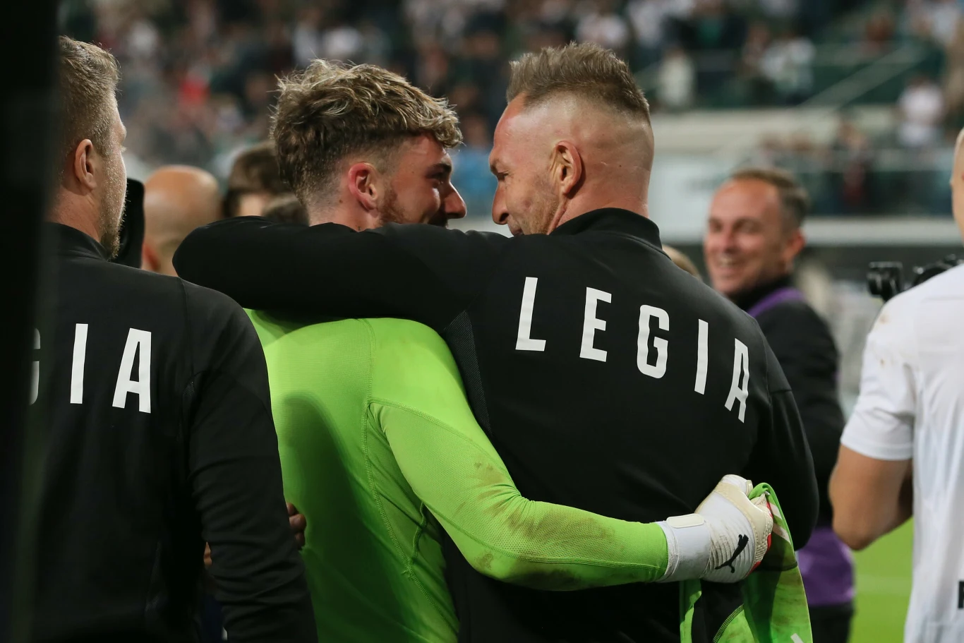 Zawodnicy drużyny Legia Warszawa w objęciu świętujący na stadionie, otoczeni przez innych członków zespołu i ludzi w tle, ogólna atmosfera radości po meczu.