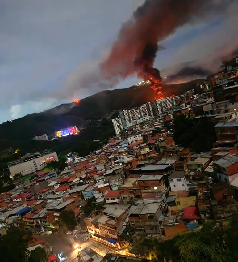 Rozległy pożar na wzgórzu w pobliżu gęsto zabudowanej dzielnicy z niskimi budynkami mieszkalnymi, gęsty dym unosi się nad płonącym obszarem, a w tle widoczne są migoczące światełka innych budynków.