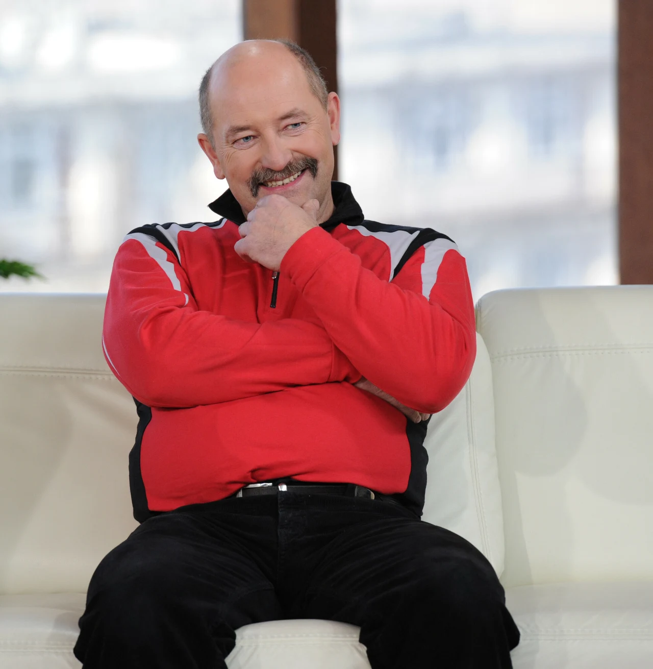 Mężczyzna w średnim wieku siedzący na białej sofie z założonymi rękoma, ubrany w czerwono-czarną bluzę sportową, uśmiechnięty, z brodą i łysiejący, w jasnym, nowoczesnym wnętrzu.