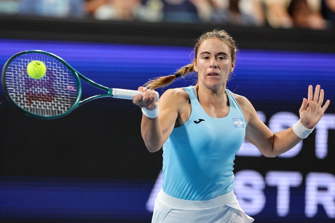 Zawodniczka w niebieskiej koszulce wykonuje forehand podczas meczu tenisowego, trzymając rakietę i koncentrując się na piłce znajdującej się tuż przed nią, w tle widoczny rozmazany napis i publiczność.