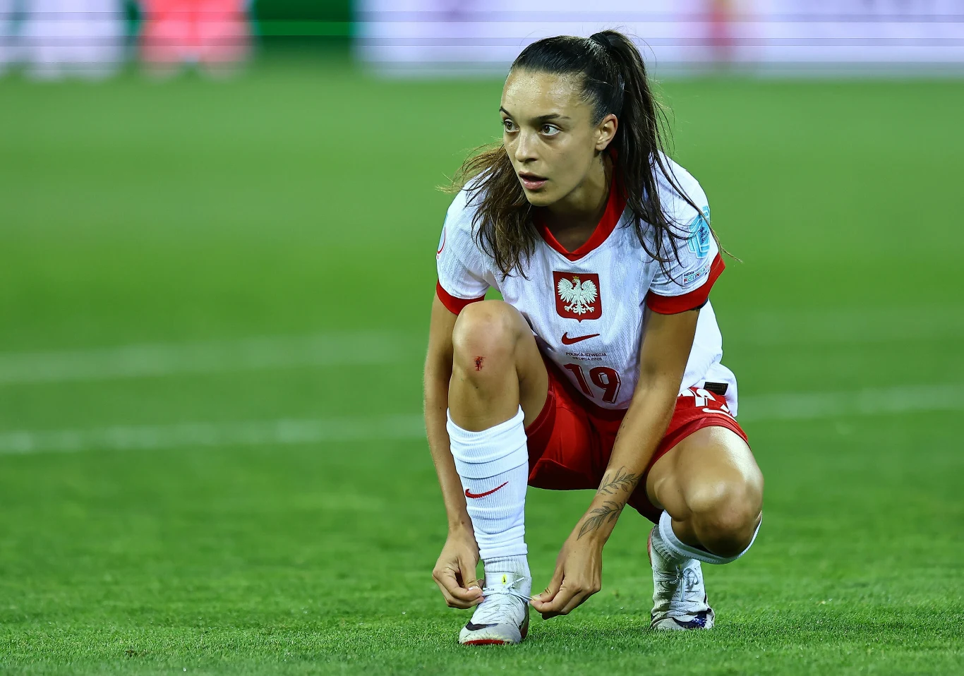 Natalia Padilla-Bidas Zawodniczka piłkarskiej reprezentacji Polski w białym stroju z czerwonymi akcentami kuca na murawie boiska i wiąże sznurowadło. Na jej kolanie widoczna jest niewielka kontuzja. W tle rozmazane światła stadionu.