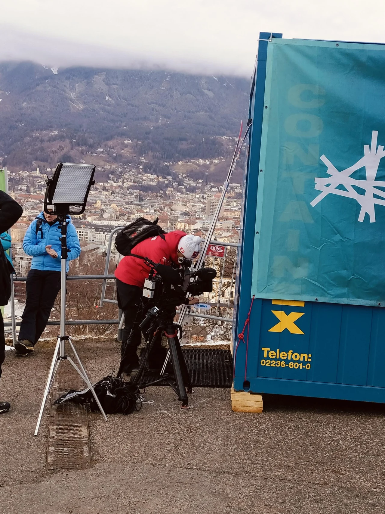 Operator kamery ustawiający profesjonalny sprzęt filmowy obok niebieskiego kontenera, w tle panorama miasta i górskie krajobrazy, przy nim osoba w niebieskiej kurtce z mikrofonem.