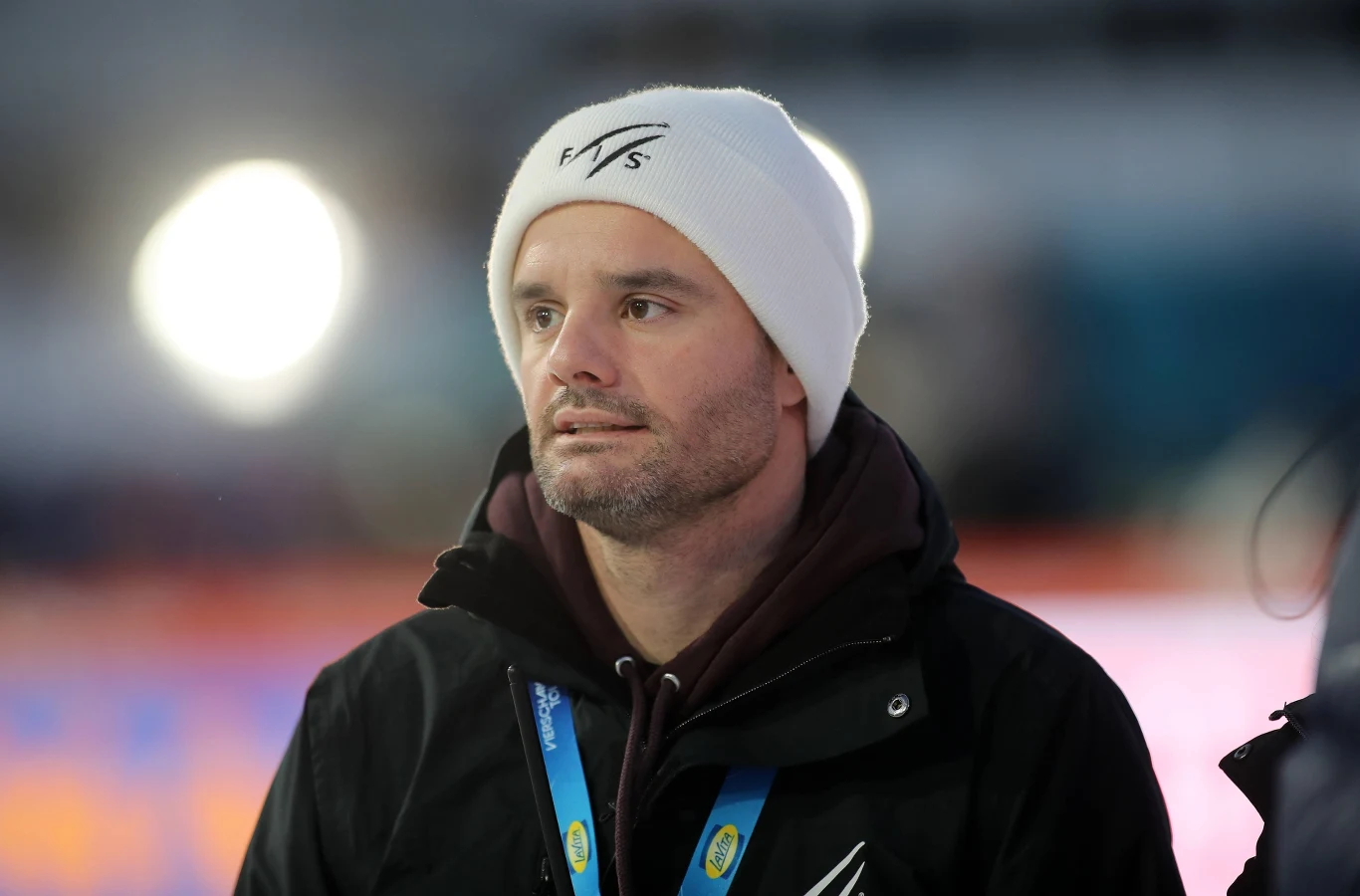 Mężczyzna z zarostem w białej czapce i czarnej kurtce sportowej z akredytacją na szyi, stojący na tle rozmytego stadionu ze źródłami światła w tle.