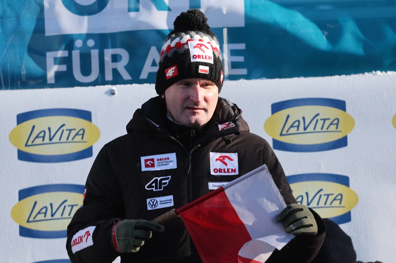 Mężczyzna ubrany w zimową kurtkę i czapkę z logo sponsorów trzyma polską flagę na tle reklamowych banerów LaVita w zimowej scenerii.