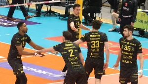 Zawodnicy drużyny siatkarskiej w czarnych strojach z żółtymi napisami ustawiają się i komunikują na boisku podczas meczu halowego.