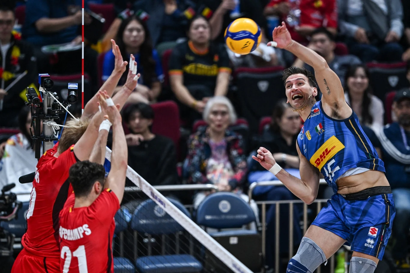 Siatkarz w niebieskim stroju wykonuje atak nad siatką, starając się zdobyć punkt, podczas gdy dwóch zawodników w czerwonych koszulkach próbuje zablokować piłkę. W tle widoczna publiczność uważnie obserwuje przebieg meczu.