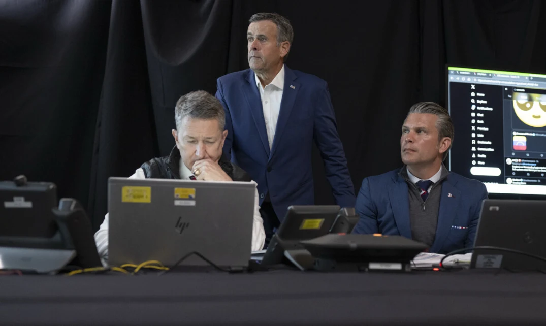 Dyrektor CIA John Ratcliffe - na zdj. w środku Trzech mężczyzn w garniturach i koszulach siedzi oraz stoi przy stole z laptopami i innym sprzętem elektronicznym, skupieni na ekranach oraz monitorze, który widoczny jest w tle.