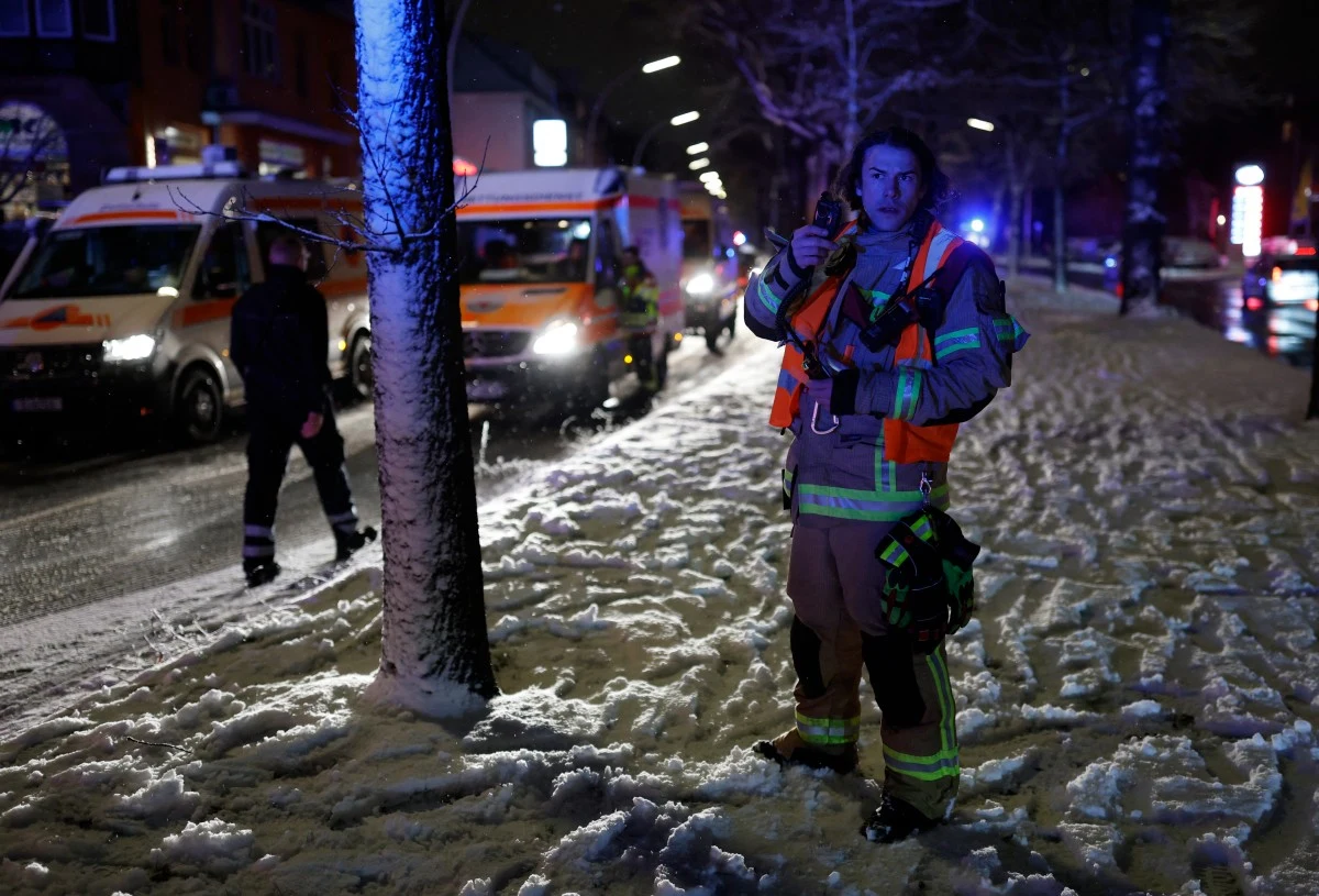Działania służb podczas awarii energetycznej w Berlinie Strażak w kamizelce odblaskowej stojący na zaśnieżonym chodniku, w tle dwa pojazdy ratunkowe z włączonymi światłami sygnalizacyjnymi oraz drugi ratownik.