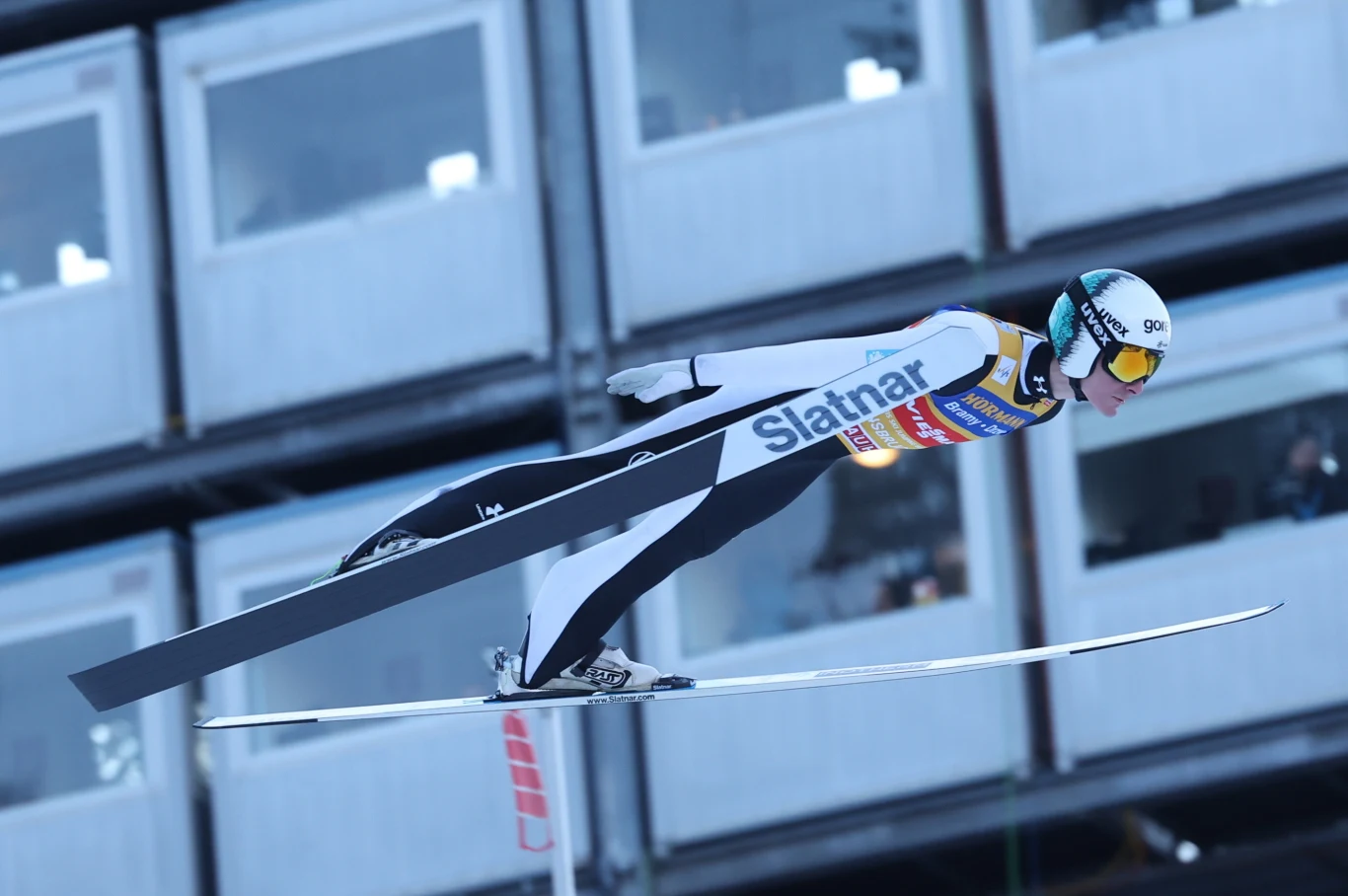 Skoczek narciarski w locie, ubrany w profesjonalny kombinezon i kask, na tle budynku z oknami obserwacyjnymi. Postawa sportowca dynamicznie oddaje moment skoku.