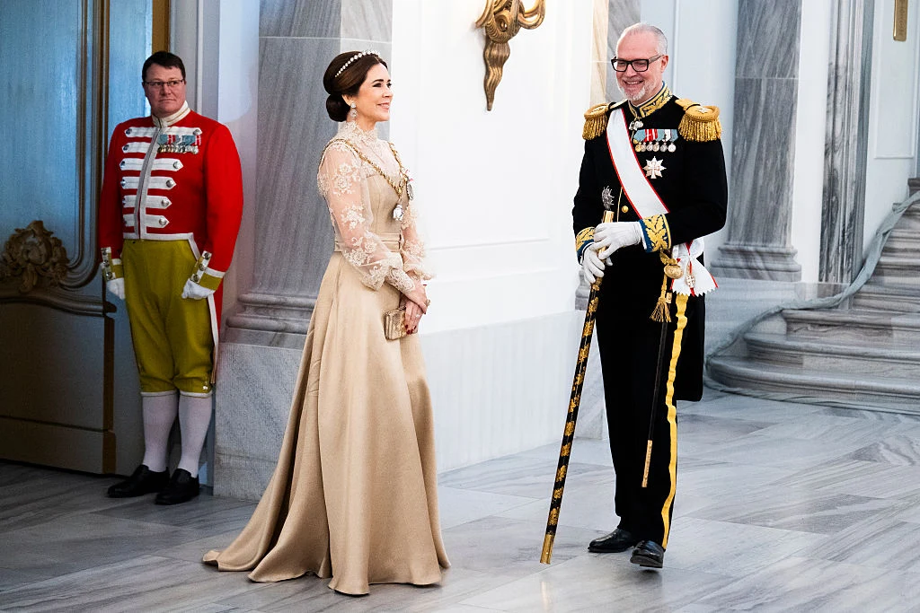 Trzy osoby w eleganckich, uroczystych strojach znajdują się w jasnej, marmurowej sali. Kobieta w długiej, beżowej sukni oraz mężczyzna w czarnym mundurze z odznaczeniami i laską stoją naprzeciwko siebie. W tle mężczyzna w czerwonym mundurze pełni funkc...