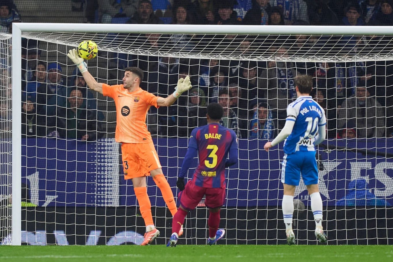 Interwencja Joana Garcii w meczu z Espanyolem Bramkarz w pomarańczowym stroju wyskakuje, próbując złapać piłkę lecącą w stronę bramki. Obok stoi piłkarz w bordowo-niebieskim stroju z numerem 3 i piłkarz drużyny przeciwnej z numerem 11 na plecach. W tle widoczni kibice na trybunach.