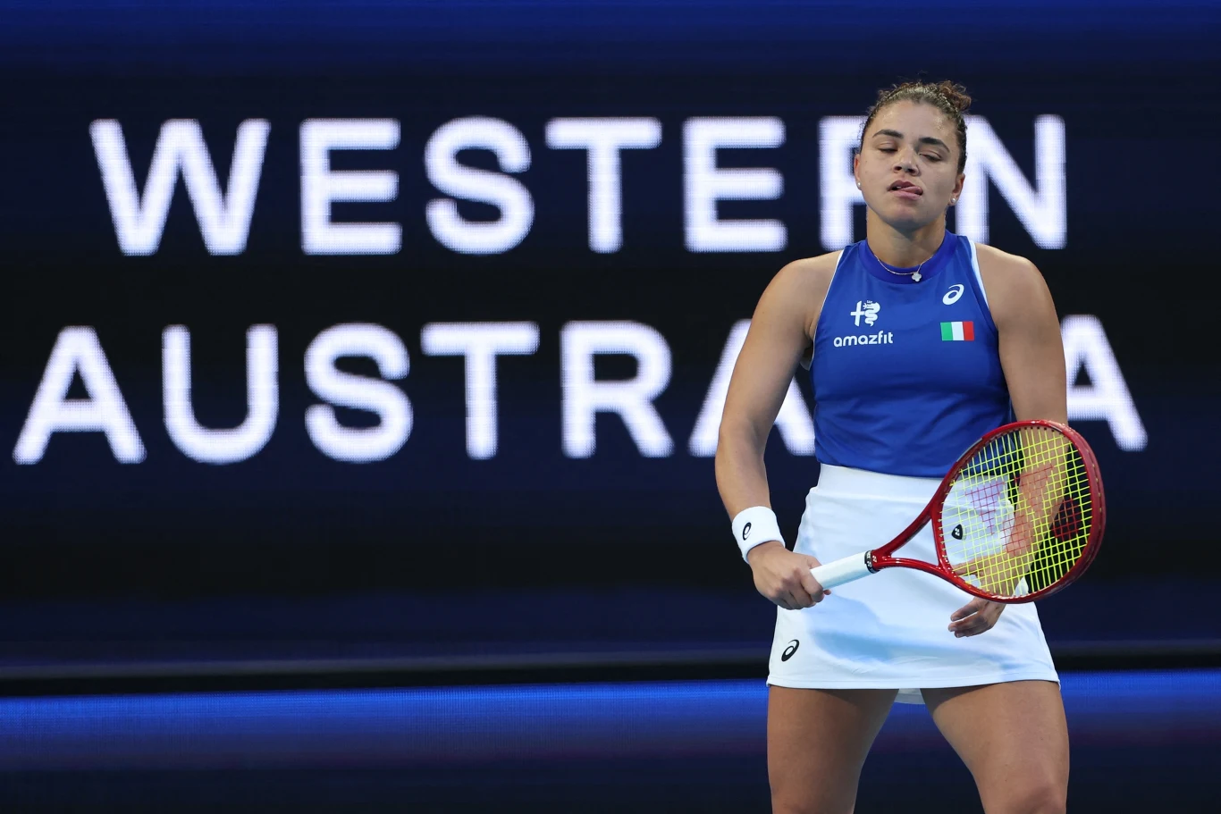 Tenisistka w niebieskiej koszulce i białej spódniczce z rakietą w dłoni stoi na korcie, zamykając oczy i wyrażając emocje. Za nią w tle widoczny jest napis Western Australia.