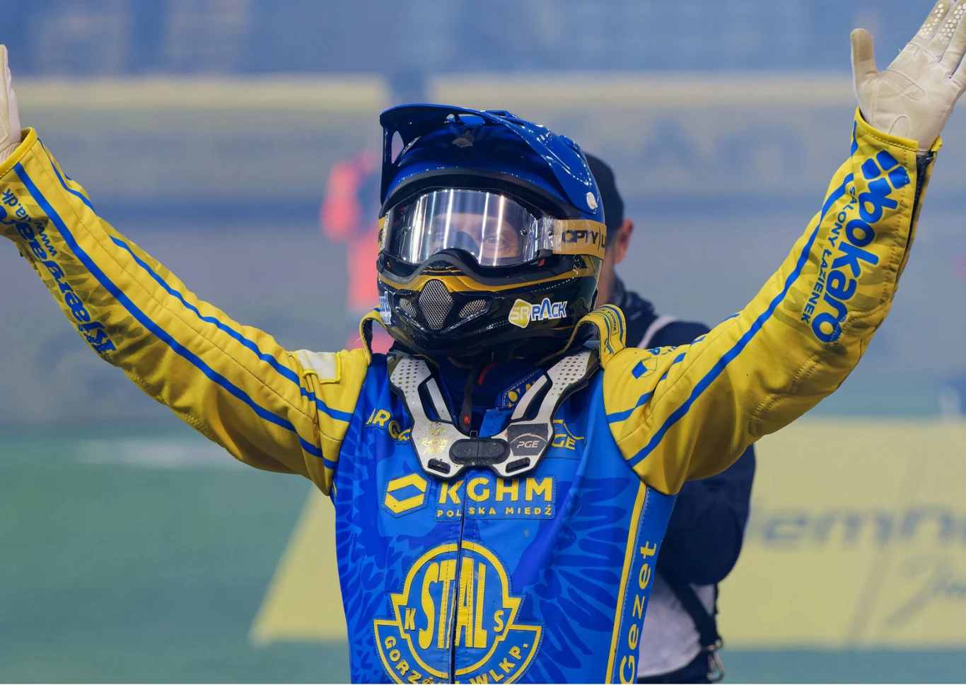 Zawodnik żużlowy w niebiesko-żółtym kombinezonie z logo klubu Stal Gorzów Wielkopolski stoi z uniesionymi rękami w geście triumfu, mając na głowie kask i gogle ochronne, w tle rozmazane sylwetki innych osób oraz tor żużlowy.