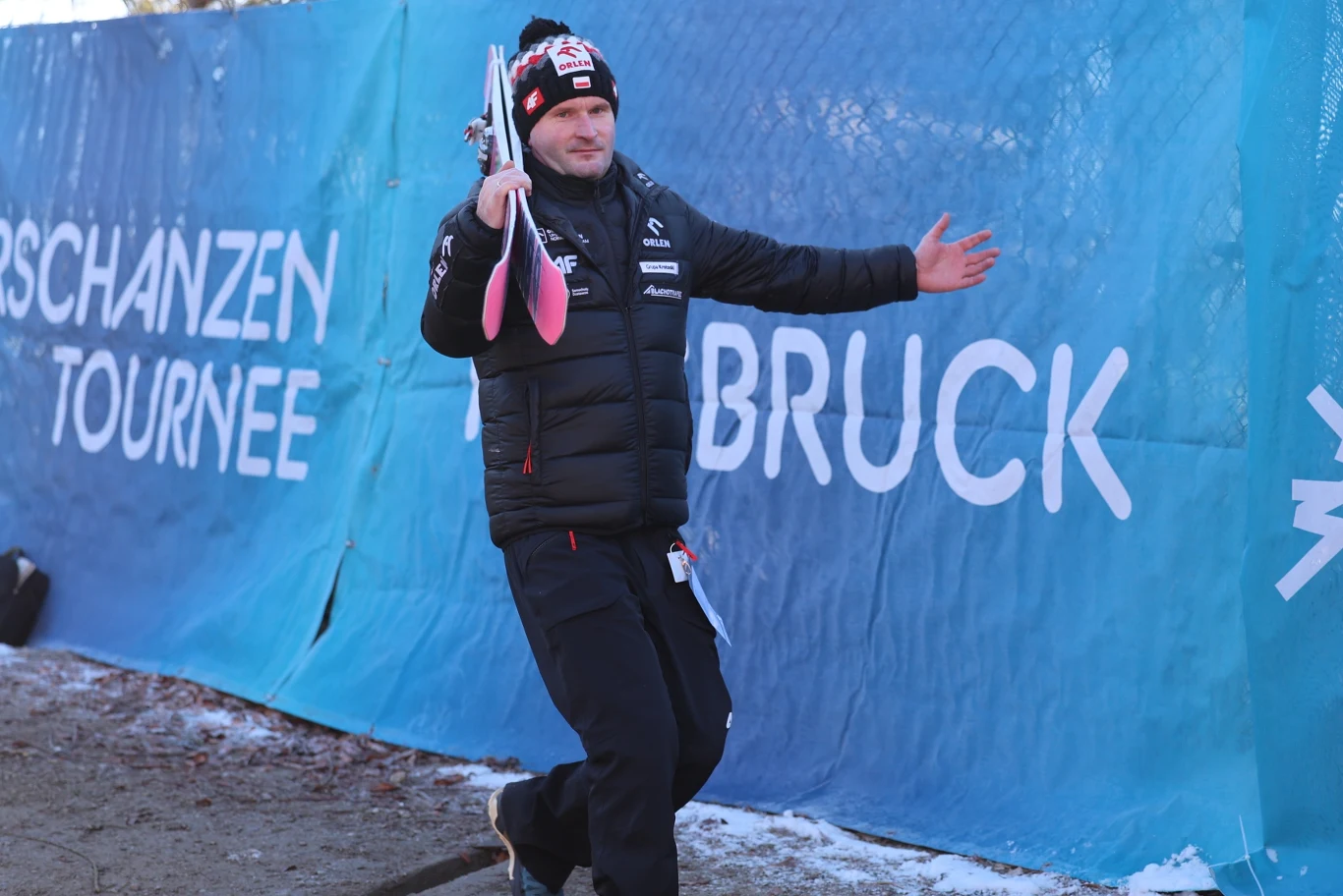 Mężczyzna ubrany w sportową kurtkę i czapkę z logo, trzymający narty i rozkładający szeroko ręce przed niebieskim banerem z napisem 'SCHANZEN TOURNEE INNSBRUCK'. Tło tworzy ogrodzenie i fragment ziemi pokrytej resztkami śniegu.