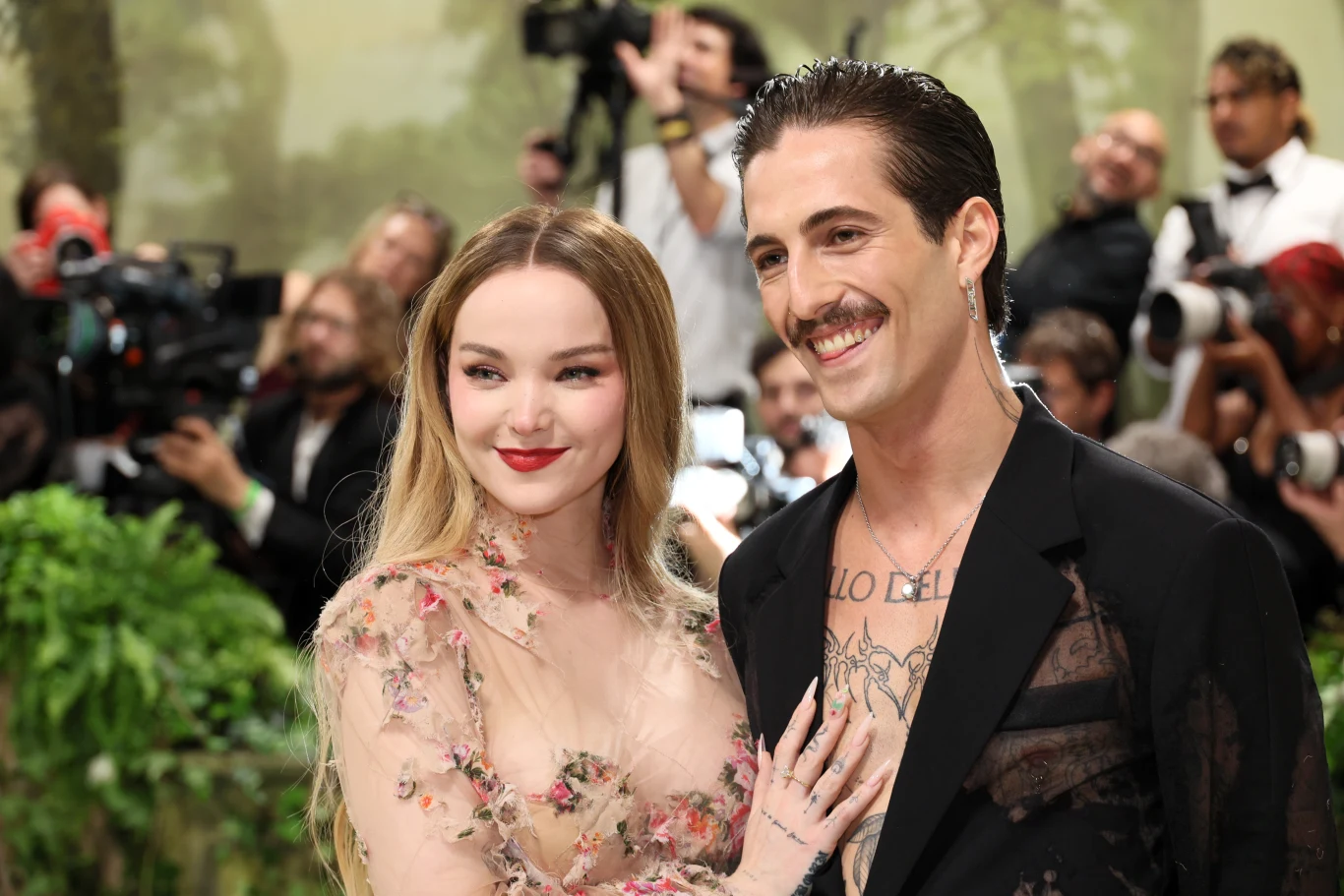 Dove Cameron i Damiano David ogłosili zaręczyny Uśmiechnięta para ubrana w eleganckie stroje, kobieta w jasnej, ozdobnej sukni, mężczyzna w czarnym garniturze bez koszuli pod spodem i tatuażami na klatce piersiowej, w tle fotografowie i zieleń dekoracyjna.