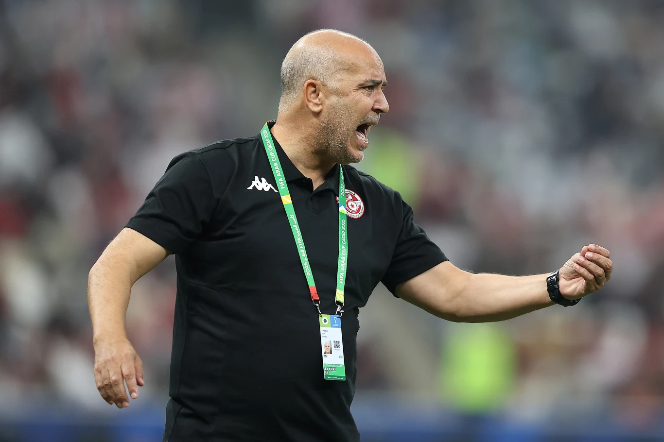 Łysy mężczyzna w czarnej koszulce z logo drużyny narodowej gestykuluje energicznie, trzymając dłoń zaciśniętą w pięść. Na szyi zawieszony identyfikator oraz zielona smycz, w tle rozmazane trybuny stadionu.