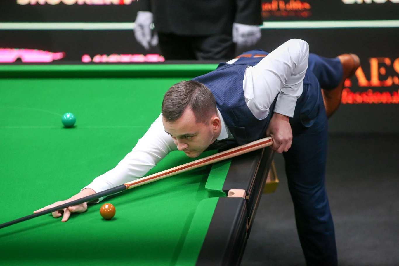 Zawodnik wykonuje uderzenie przy stole bilardowym w trakcie meczu snookera, skupiony na precyzyjnym poprowadzeniu kija, w tle widoczne elementy sędziowskie.