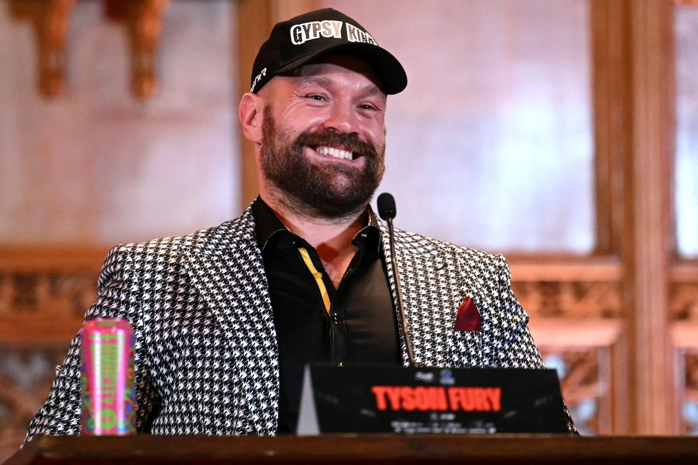 Uśmiechnięty mężczyzna w czapce z daszkiem i wzorzystej marynarce stoi za mikrofonem, przed nim znajduje się tabliczka z napisem Tyson Fury oraz kolorowa puszka napoju.