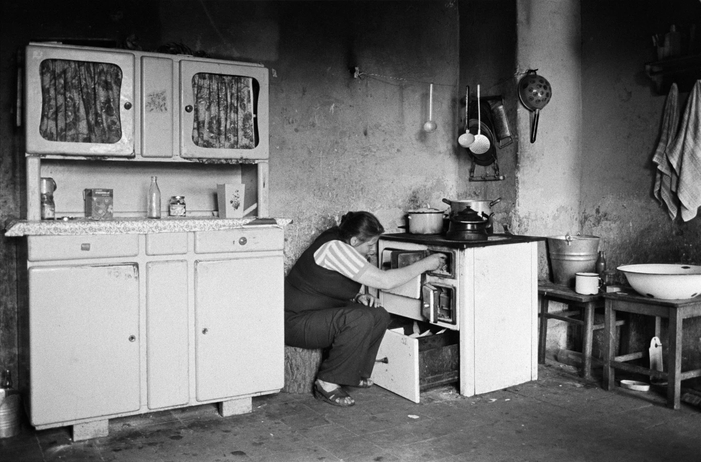 Kobieta w kuchni z dawnych lat, klęcząca przy piecu i zaglądająca do jego wnętrza. Po lewej stronie stara szafka kuchenna, na ścianie wiszą przybory kuchenne i ręcznik, obok widoczna miska na stole. Ściany noszą ślady zużycia i patyny.