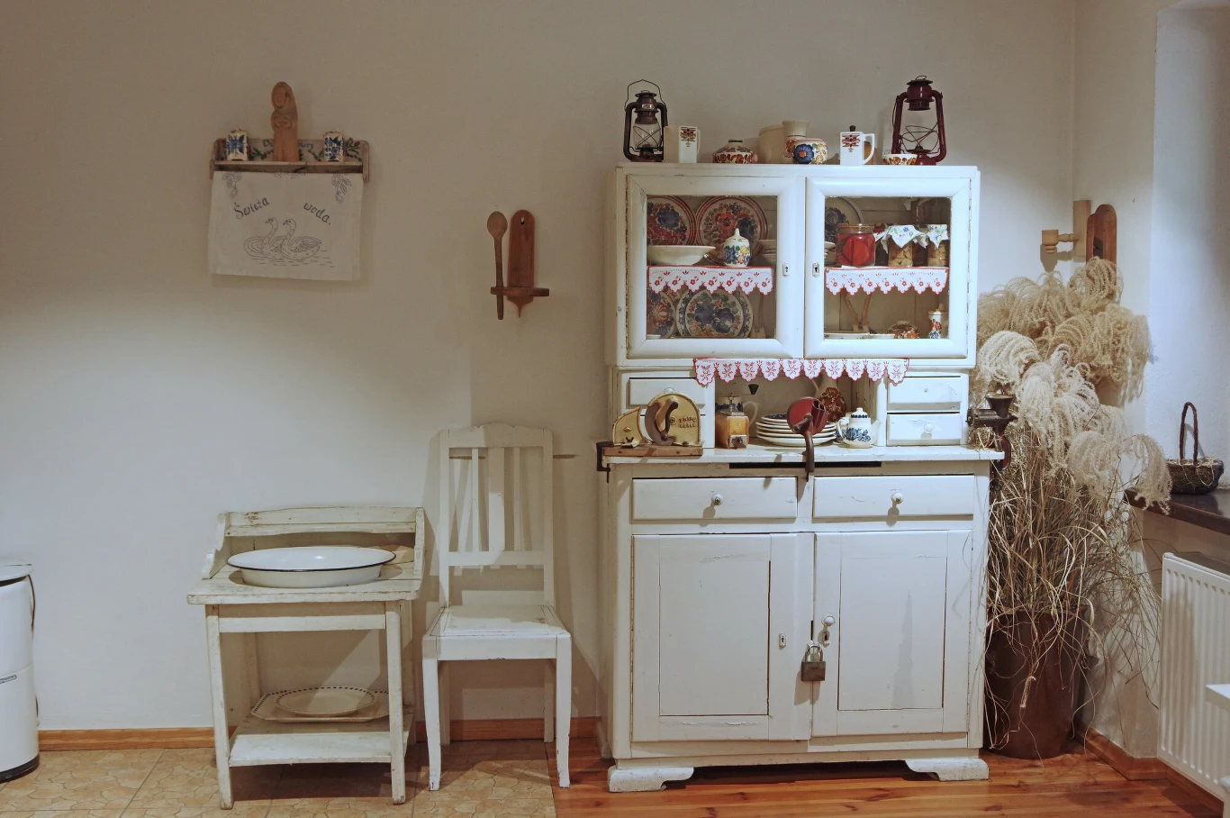 Biała drewniana szafka kuchennego kredensu z ozdobnymi serwetkami i starodawną zastawą, obok stoją białe krzesło, mała myjnia z miską oraz dekoracje w postaci suszonych traw ustawionych przy oknie.