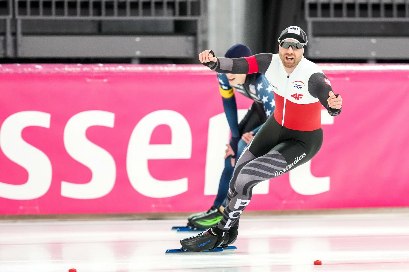 Zawodnik w biało-czerwonym stroju podczas wyścigu na łyżwach szybkich na torze lodowym, za nim widoczny drugi zawodnik w niebieskim stroju; w tle różowa reklama i trybuny.