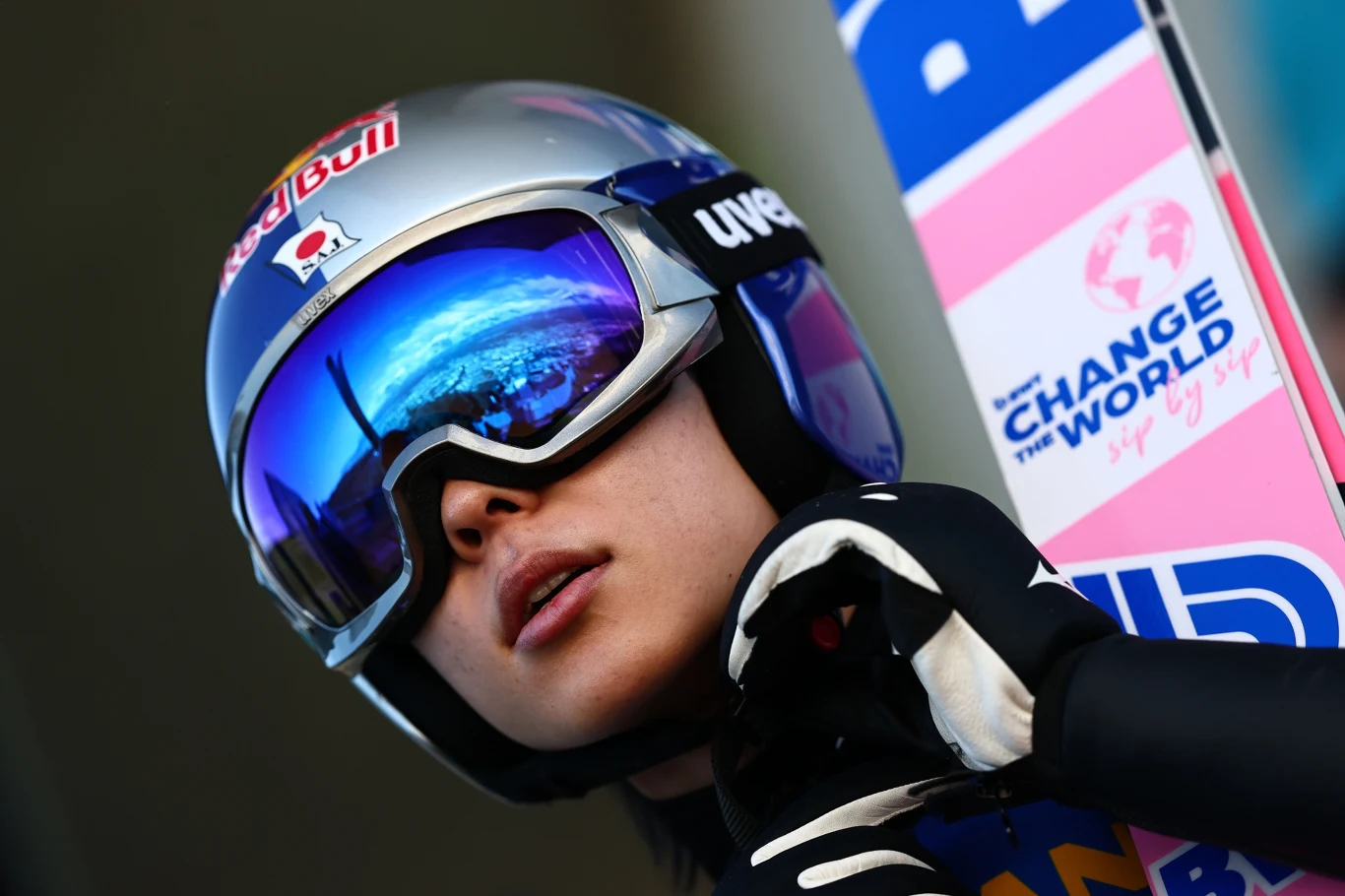 Sportowiec w kasku i goglach narciarskich przygotowuje się do skoku narciarskiego, w odbiciu gogli widoczny fragment skoczni i niebo, obok narty z napisem "Change the World".