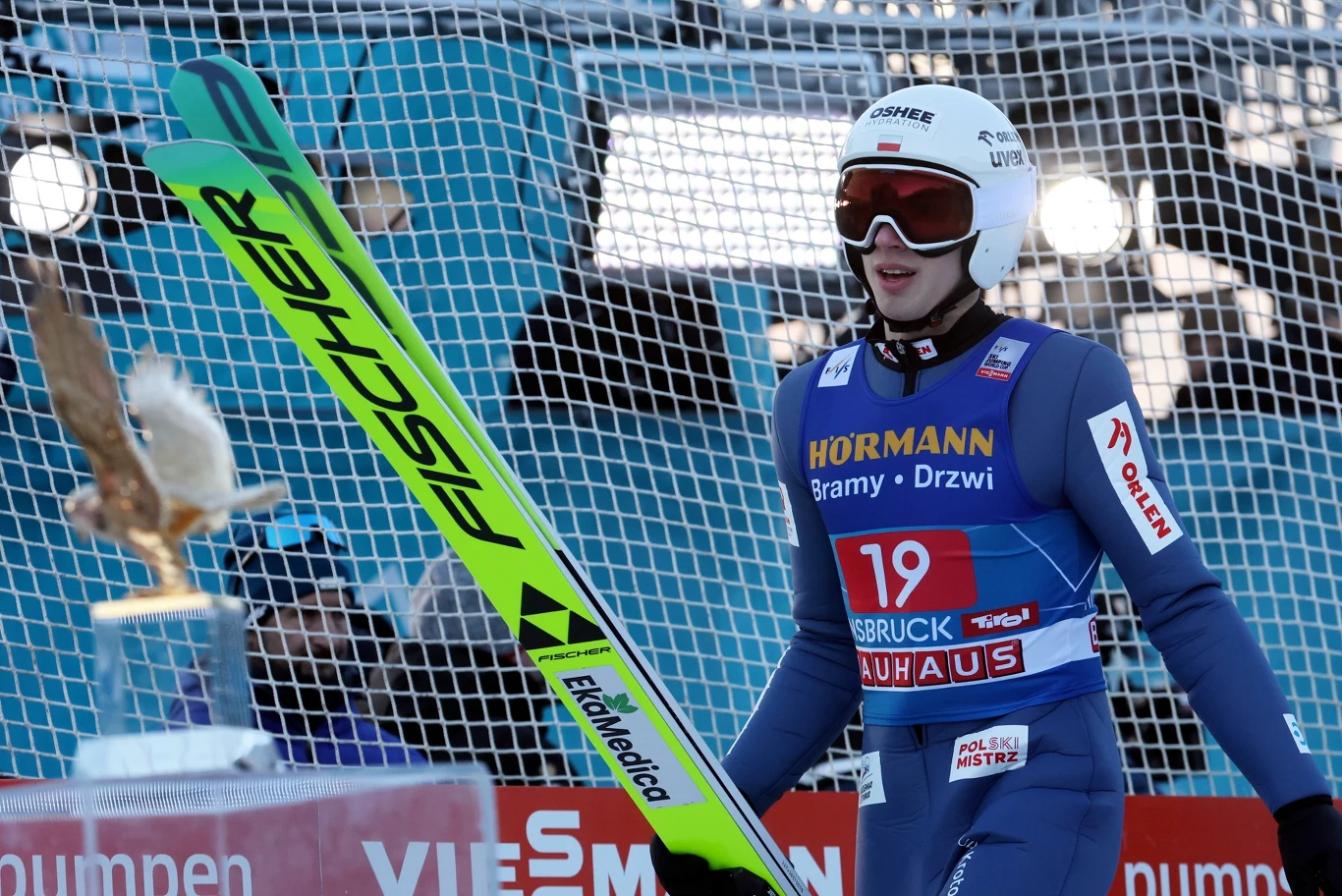 Skoczek narciarski w kombinezonie startowym idący z nartami po zakończonym skoku, w tle siatka ochronna oraz elementy infrastruktury skoczni narciarskiej.