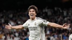 Piłkarz w białym stroju drużyny Real Madryt z szeroko rozpostartymi ramionami świętuje zdobycie gola na tle rozmytej publiczności.