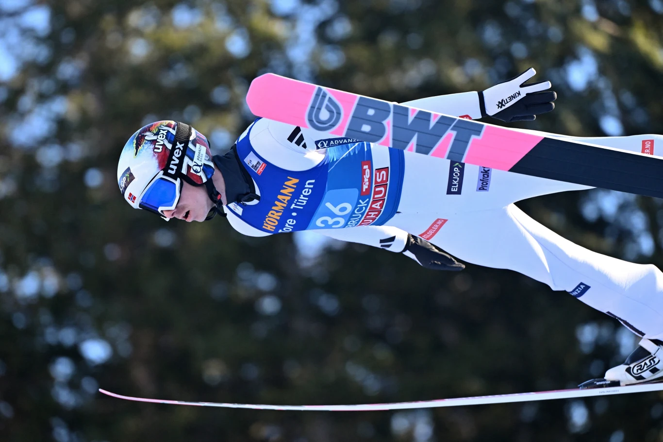 Halvor Egner Granerud Skoczek narciarski w locie w pełnym ekwipunku z numerem startowym, kaskiem i goglami, na tle rozmytych drzew, dynamiczna poza podczas konkursu sportowego.