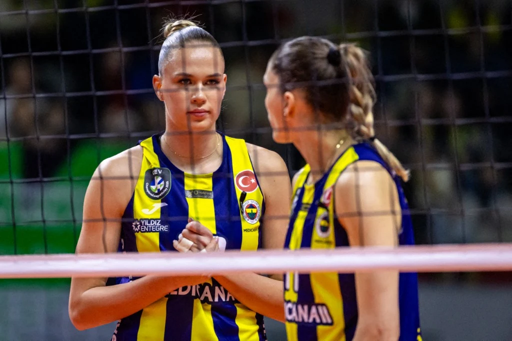 Dwie siatkarki w żółto-niebieskich strojach drużynowych stojące po tej samej stronie siatki, prowadzą rozmowę podczas meczu siatkówki w hali sportowej.