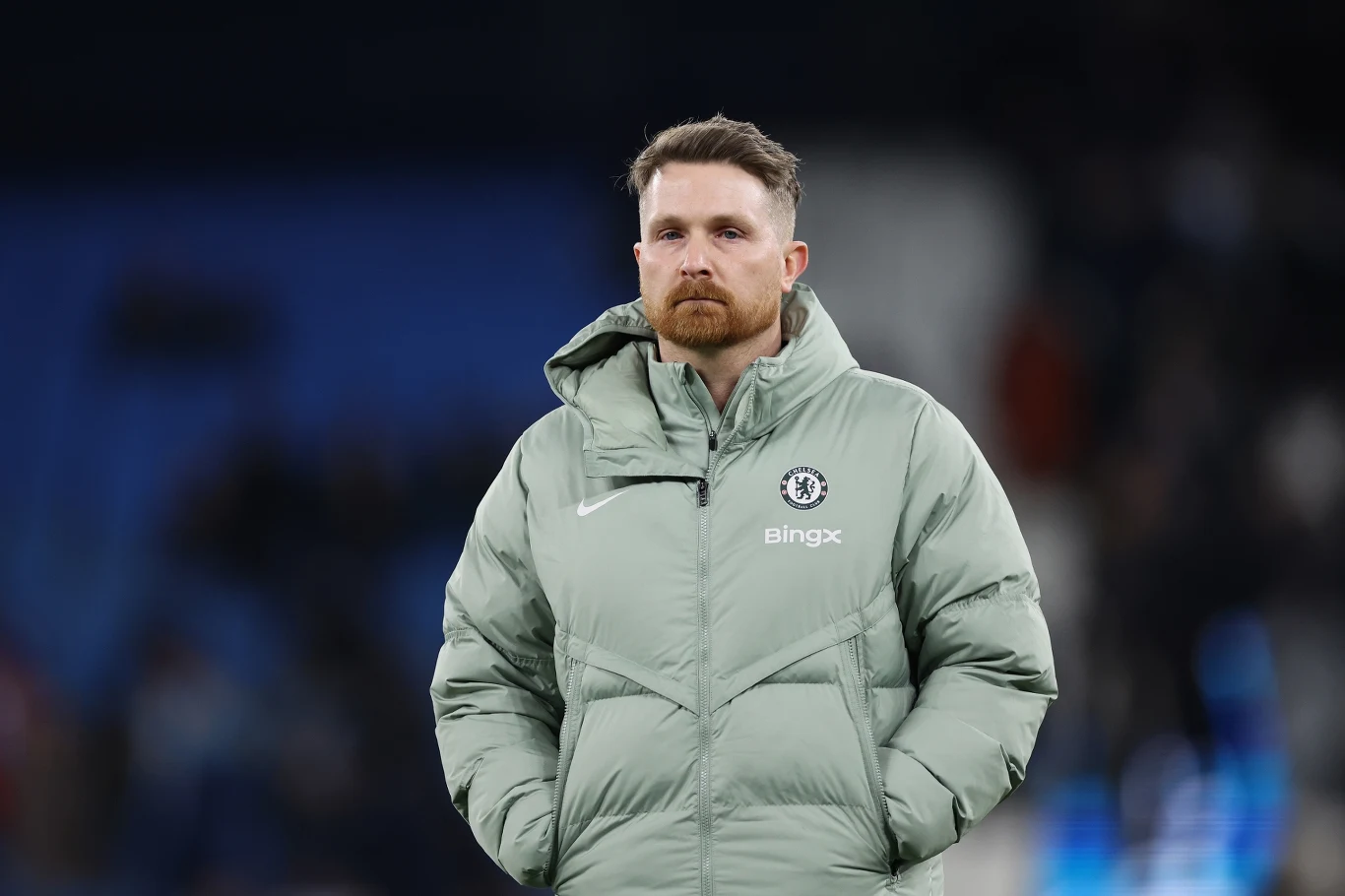 Mężczyzna w jasnej kurtce sportowej z logo Chelsea i BingX stoi na stadionie, tło jest rozmyte, skupiając uwagę na jego wyrazie twarzy i stroju.