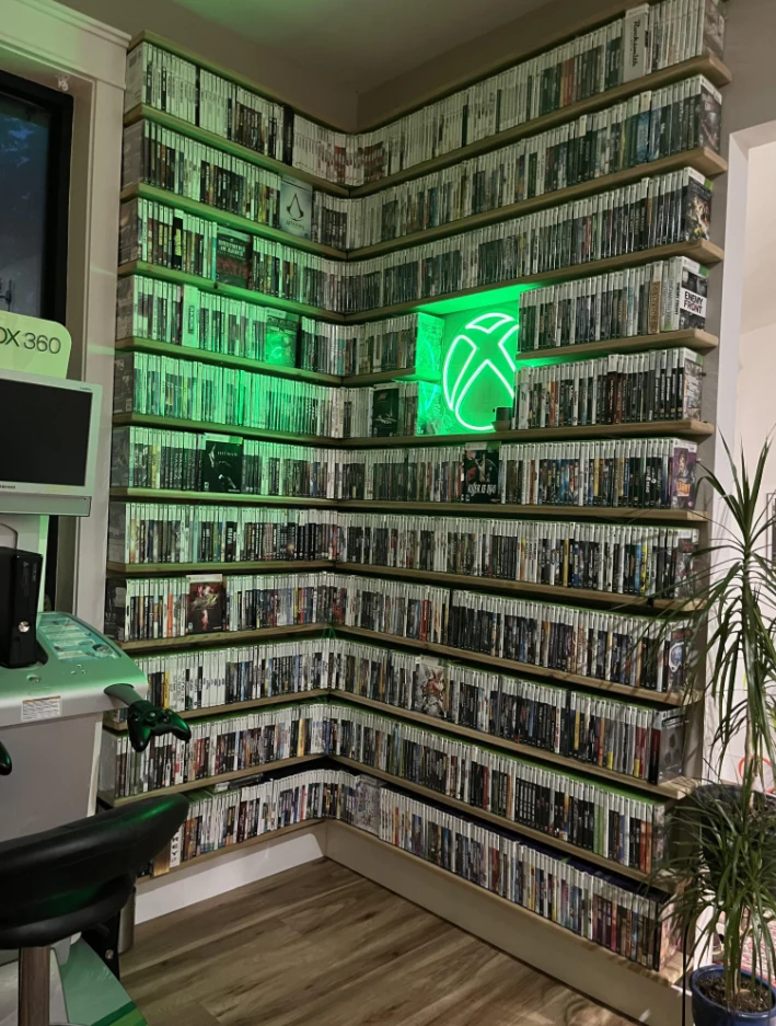 Ściana z półkami pełnymi gier na konsolę Xbox 360, podświetlona zielonym światłem z logo Xbox, w pobliżu stanowisko do gry z monitorem i padem, a także roślina doniczkowa po prawej stronie.