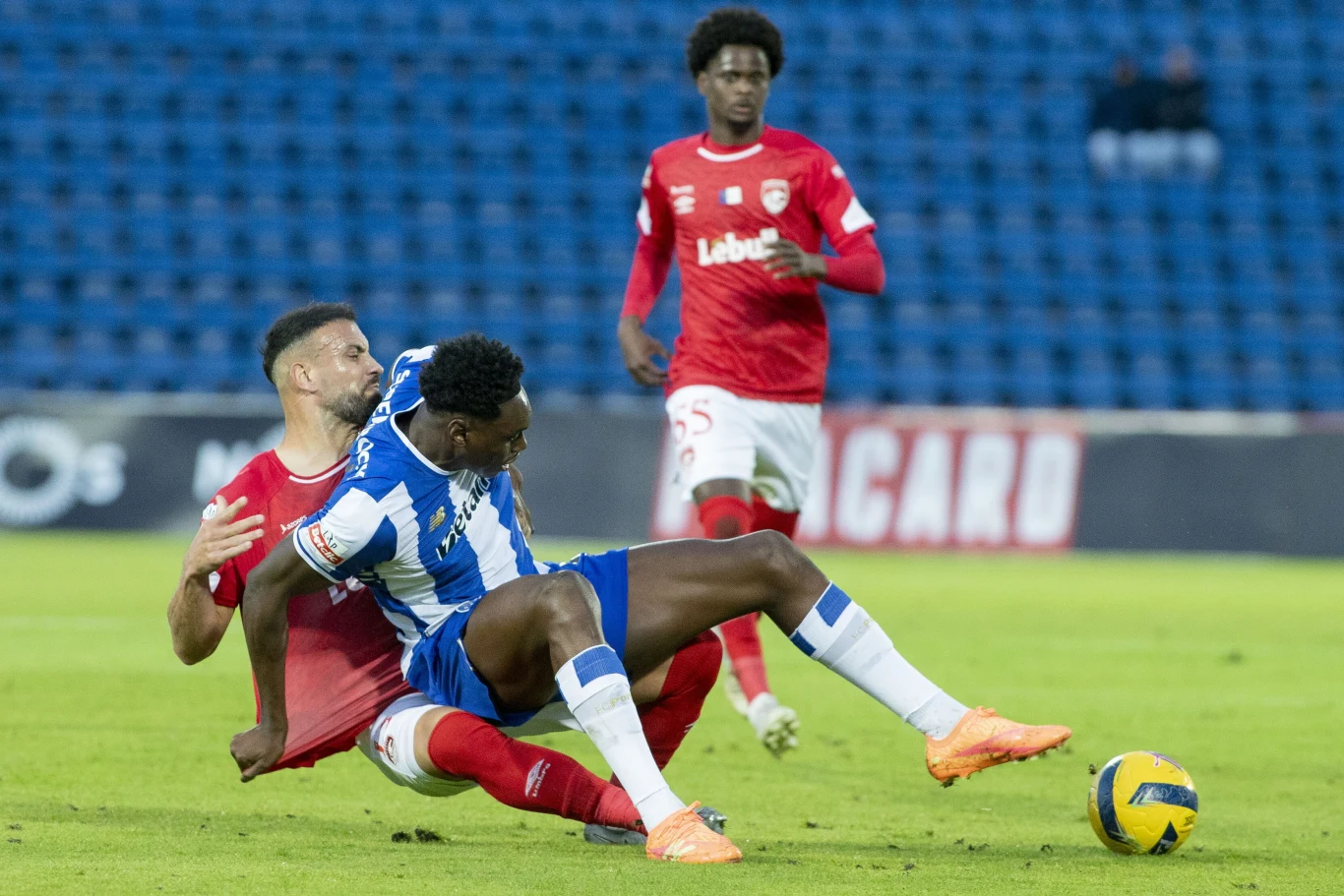 Samu (na pierwszym planie) nie miał łatwego życia z defensorami Santa Clara, ale do siatki zdołał trafić Dwóch piłkarzy rywalizuje o piłkę podczas meczu piłkarskiego; jeden z nich w niebiesko-białym stroju jest powalony na murawę przez przeciwnika w czerwonym stroju, w tle widoczny trzeci zawodnik oraz pusta trybuna stadionowa.