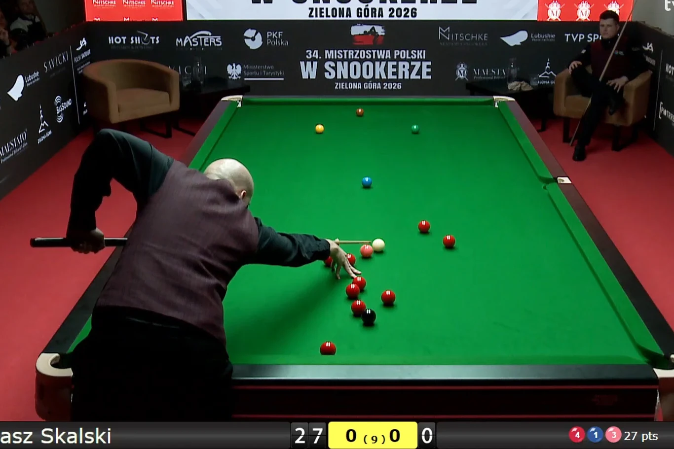 Zawodnik grający w snookera pochylony nad zielonym stołem bilardowym wykonuje precyzyjny strzał. W tle widoczne są reklamy sponsorów oraz puste fotele przy ścianie. Licznik punktów oraz nazwisko zawodnika pojawiają się na ekranie.