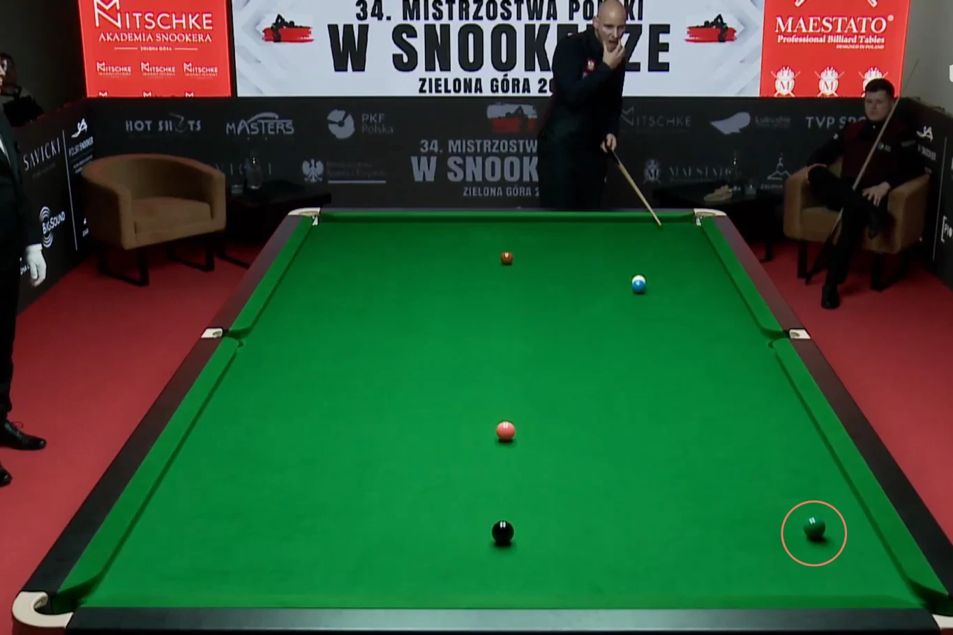 Stół bilardowy z zielonym suknem, na którym rozmieszczone są bile do gry w snookera, jeden z zawodników przygotowuje się do uderzenia kijem, widoczni są także sędziowie oraz banery reklamowe wokół stołu.