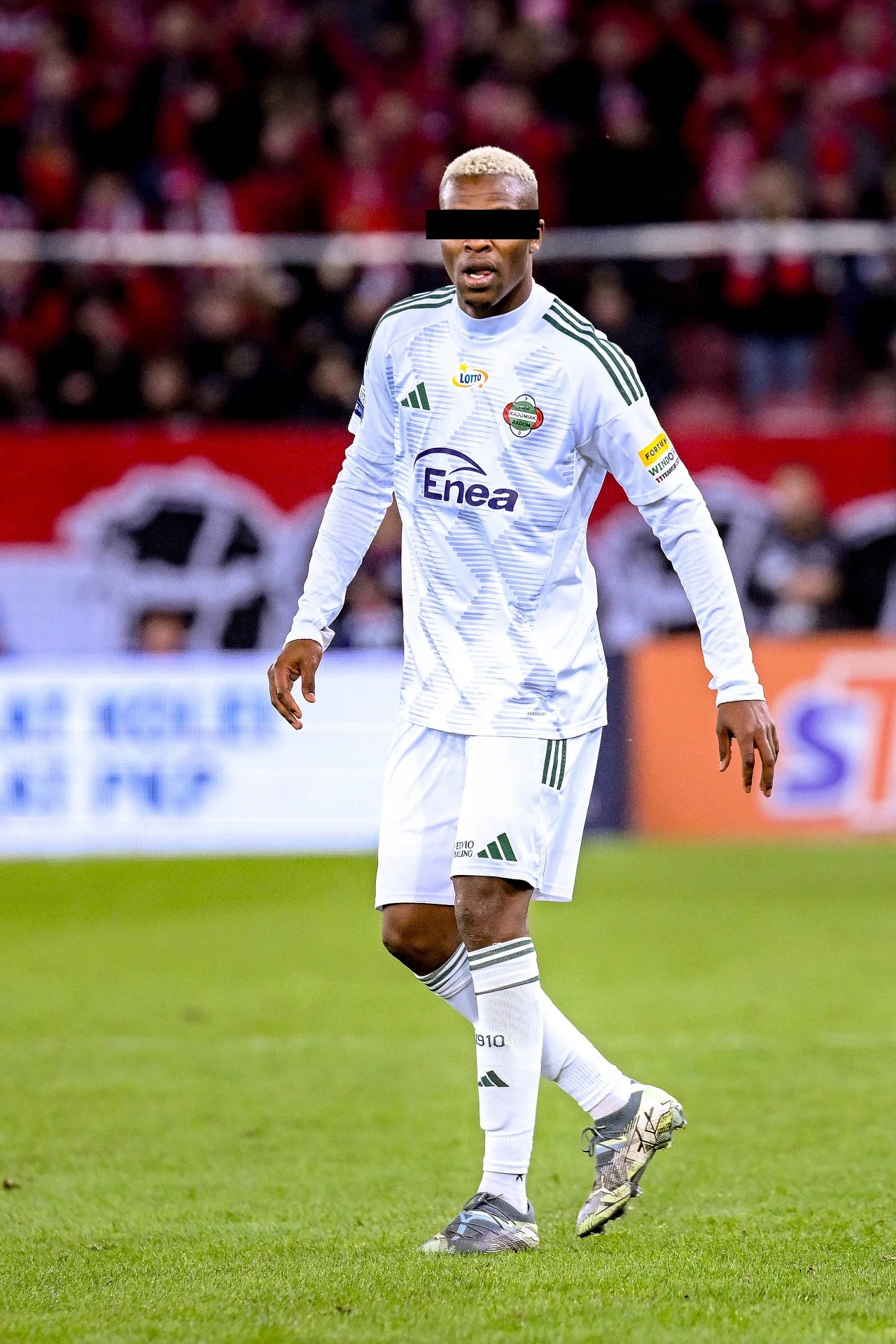 Piłkarz w białym stroju meczowym z logotypami sponsorów na tle murawy oraz trybun wypełnionych kibicami, zawodnik ma zasłonięte oczy czarnym paskiem.