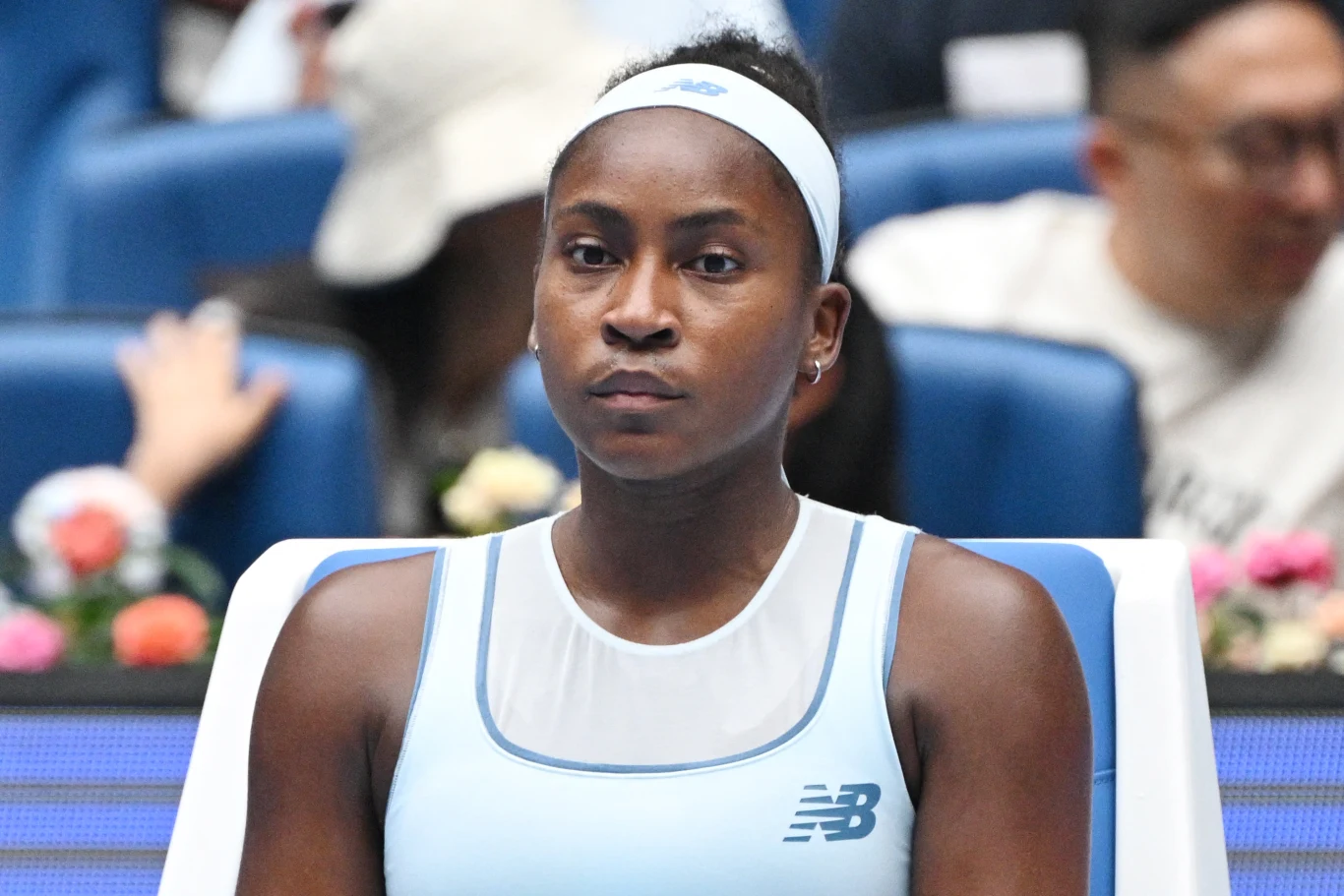 Młoda kobieta w sportowym stroju tenisowym siedzi skupiona na bocznym siedzisku kortu, w tle widoczni są kibice oraz kwiaty.