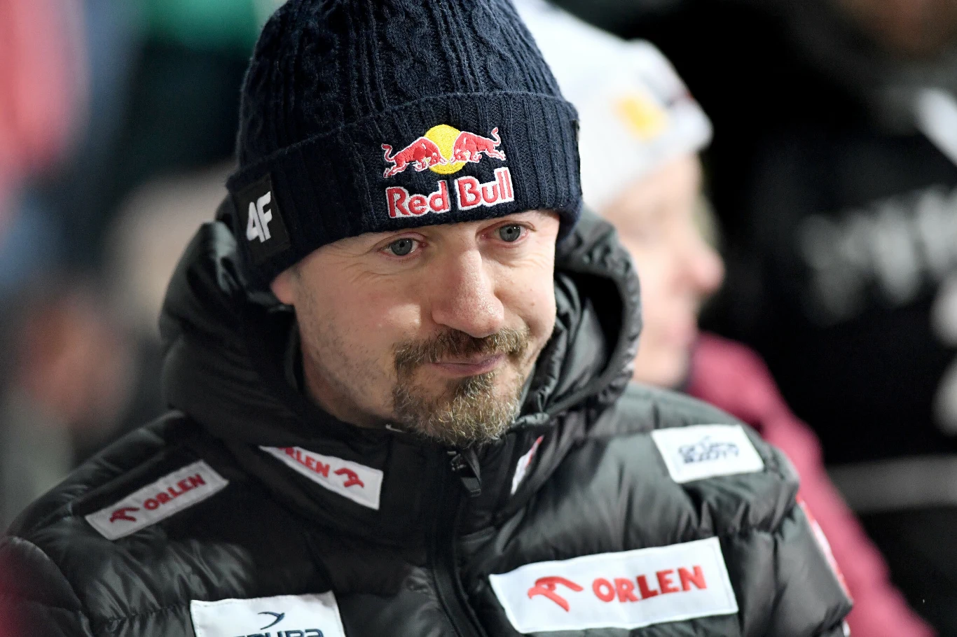 Mężczyzna ubrany w zimową kurtkę z logo Orlen i czapką z logo Red Bulla, patrzący lekko w bok ze spokojnym wyrazem twarzy, na tle rozmytych postaci w jasnych ubraniach.