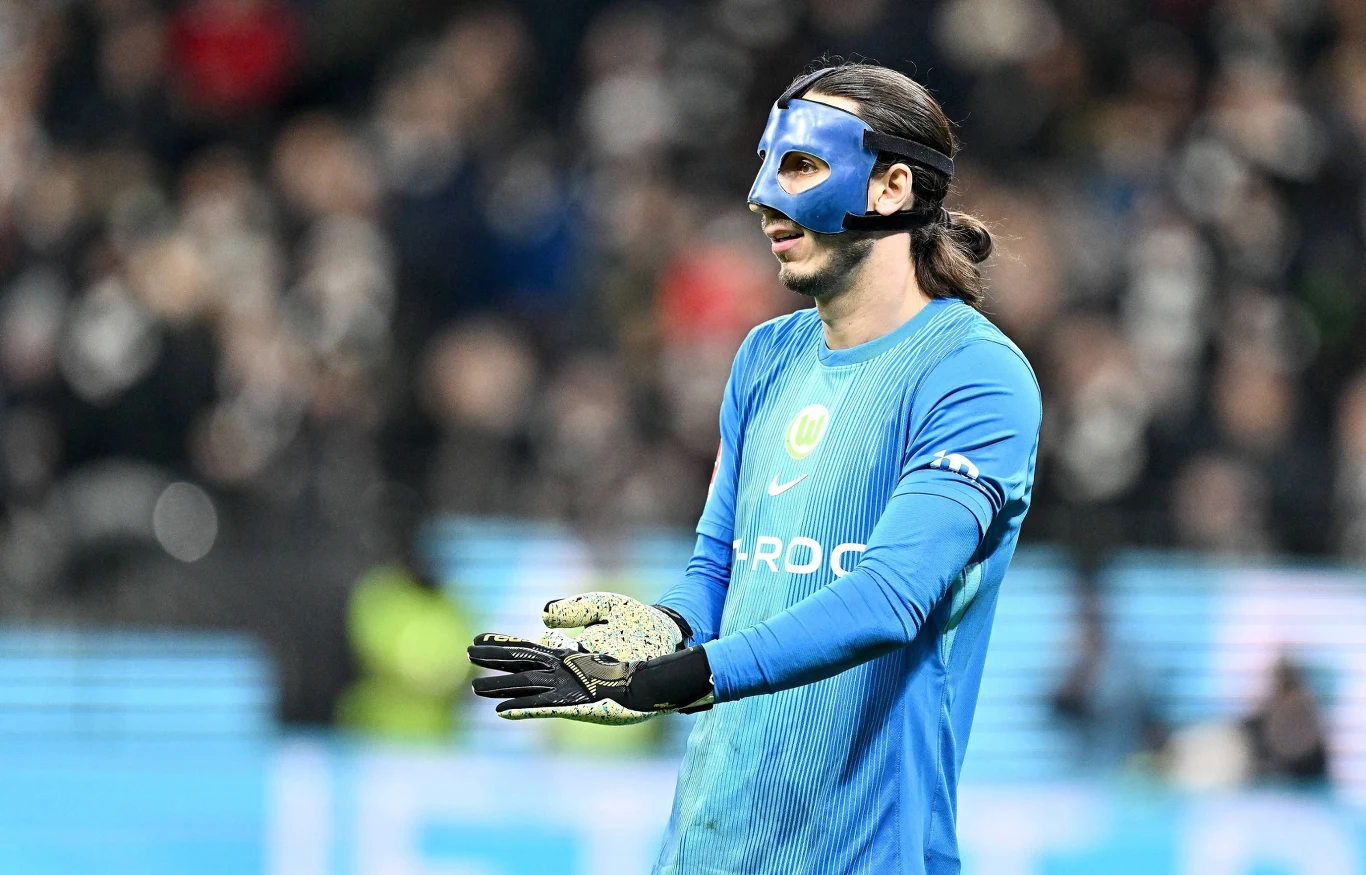 Piłkarz w niebieskim stroju bramkarskim i czarnej masce na twarzy stoi na boisku, trzyma dłonie rozłożone, w tle rozmyci kibice.