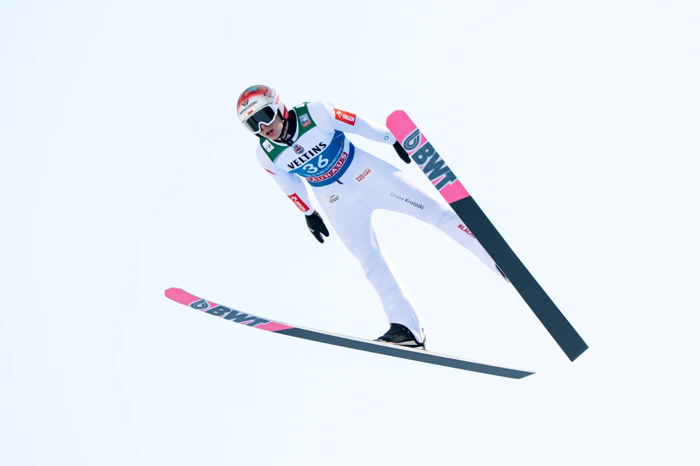 Skoczek narciarski ubrany w biały kombinezon wykonuje skok na nartach na tle jasnego nieba, widoczny sprzęt sportowy oraz numer startowy zawodnika.