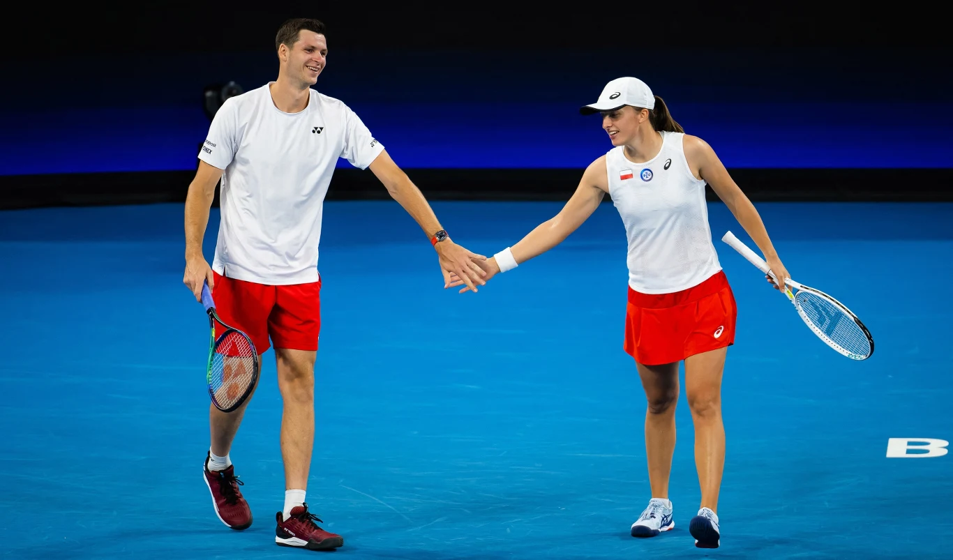 Dwójka tenisistów w sportowych strojach świętuje udaną akcję na korcie, podając sobie ręce i uśmiechając się do siebie. Tło stanowi niebieski kort tenisowy.